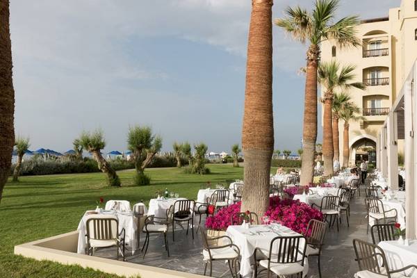Außenbereich des Aquila Rithymna Beach Hotels mit Tischen und Stühlen, umgeben von Palmen und Blumen.