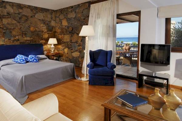 Schlafzimmer im Aquila Rithymna Beach Hotel mit Steinwänden, einem Bett, zwei Sesseln und einem Fernseher.