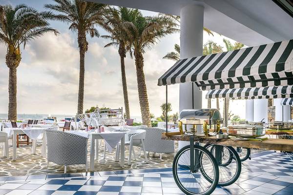 Außenbereich des Minos Imperial Beach Resorts mit Palmen, blau-weißem Boden und Tischen.
