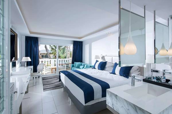 Innenansicht eines Zimmers im Minos Imperial Beach Resort mit weißem Bett, blauen Kissen und modernem Badezimmer.