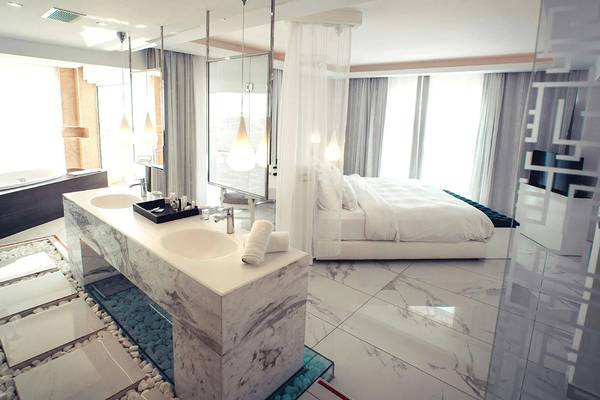Innenansicht eines Schlafzimmers im Minos Imperial Beach Resort mit Marmorbad, Doppelbett und zwei Waschbecken.