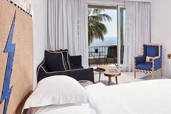 Zimmer im Royal Myconian Resort mit Balkon und Meerblick, inklusive Bett, Couch, Fernseher und Nachttisch.