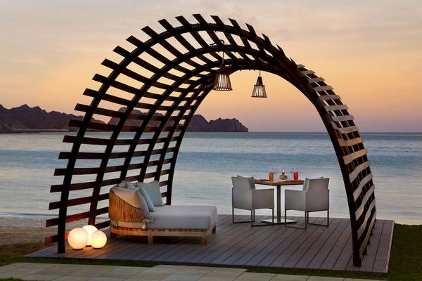 Abendessen am Strand des Al Bustan Palace, einem Ritz Carlton Hotel, mit einem Tisch, Stühlen und einer Liege, umgeben von einer bogenförmigen Struktur.