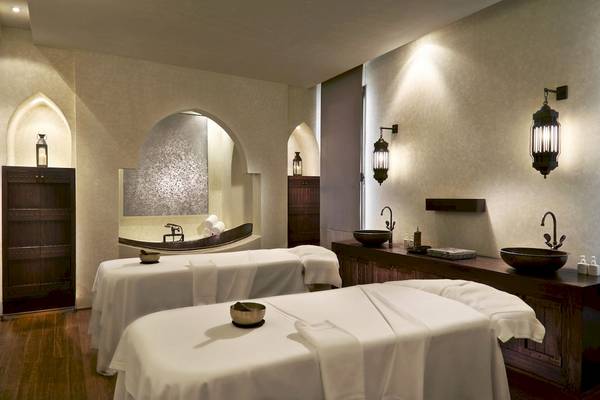 Spa-Behandlungsraum im Al Bustan Palace, zwei Behandlungstische mit weißen Laken und Kerzen, Holzboden und traditionelle Beleuchtung.