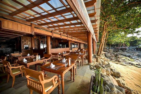 Terrassenrestaurant des Santhiya Koh Phangan Resort & Spa mit Tischen und Stühlen unter einem Holzdach, umgeben von Bäumen und Felsen am Strand