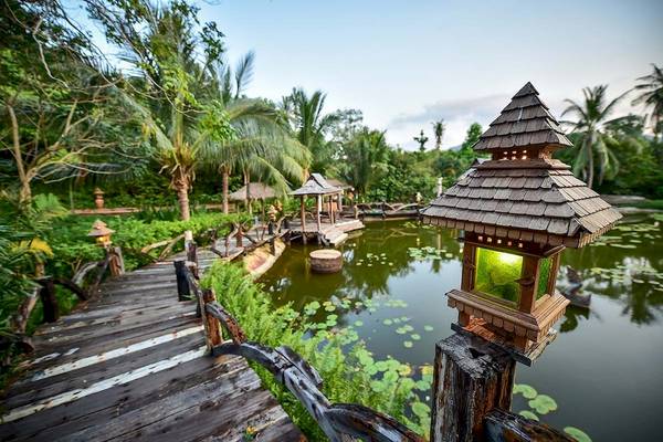 Ein Garten mit einer Holzbrücke und einem Teich, beleuchtet von Laternen, im Santhiya Koh Phangan Resort & Spa.