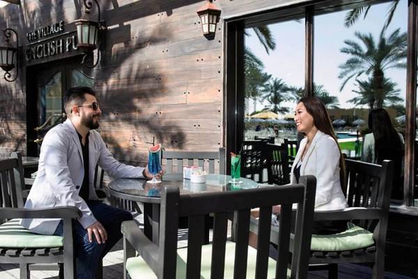 Mann und Frau sitzen vor der The Village English Pub im Al Hamra Village und unterhalten sich.