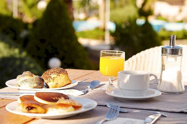 Frühstückstisch im Garten mit verschiedenen Gebäck, Orangensaft, Kaffee und Pool im Hintergrund.