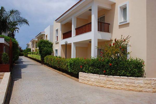 Außenansicht des Aktea Beach Village Hotels mit gepflastertem Weg und roten Blumenbüschen.