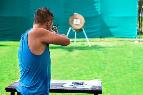 Ein Mann in einem blauen Tanktop zielt mit einer Waffe auf eine Zielscheibe auf einer Wiese.