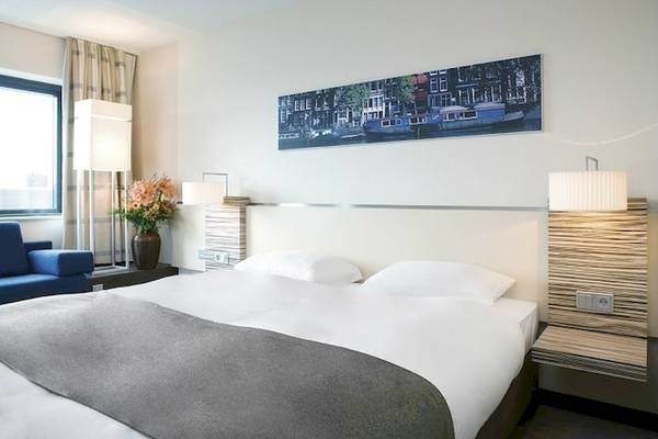 Hotelzimmer im Mövenpick Amsterdam City Centre mit Bett, Kissen, Lampe und Gemälde.