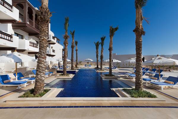 Pool-Lounge-Bereich im Ancient Sands Golf Resort mit weißen Gebäuden, blauen Liegestühlen und Sonnenschirmen.