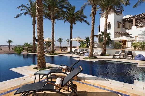 Pool-Loungebereich im Ancient Sands Golf Resort mit Sonnenliegen, Palmen und weißen Gebäuden.