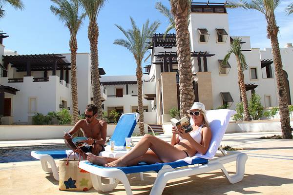 Paar am Pool im Ancient Sands Golf Resort, Frau liest Buch, Mann liest Zeitschrift, beide in Sonnenbrillen, umgeben von Palmen und Gebäuden.