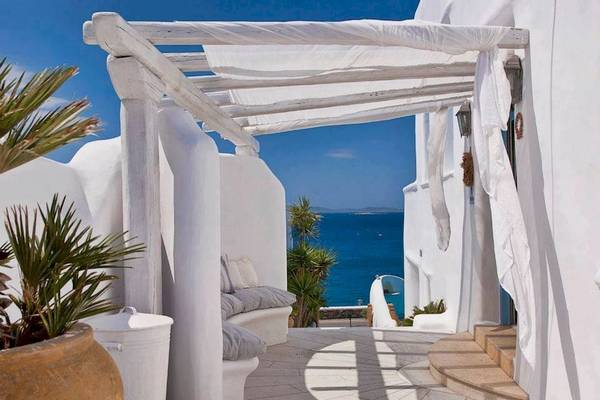 Hotelterrasse mit Meerblick, weiße Wände und Möbel, Pergola mit weißem Vorhang, Blick auf das blaue Meer.