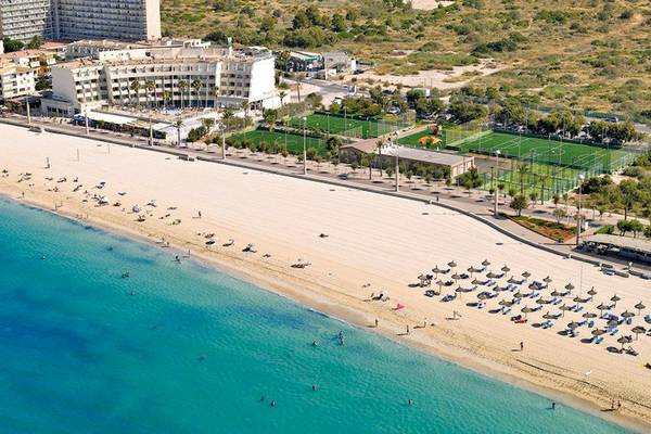 Aerial view of a beach with people, a hotel, and sports fields in the background.