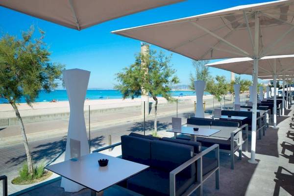 Außenterrasse im Aparthotel Fontanellas Playa mit Tischen, Stühlen und großen Schirmen, Blick auf den Strand und das Meer.