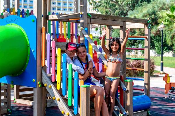 Zwei Kinder und eine Frau spielen auf einem bunten Spielplatz im Freien.