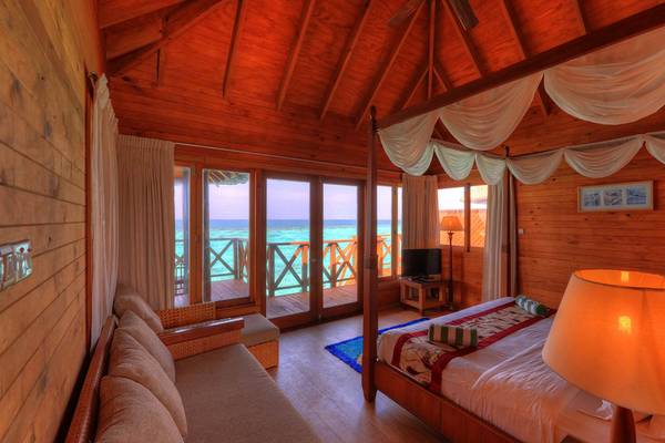Innenansicht eines Zimmers im Fihalhohi Island Resort mit Holzwänden, einem großen Bett, einem Fernseher und einem Ausblick auf das Meer.