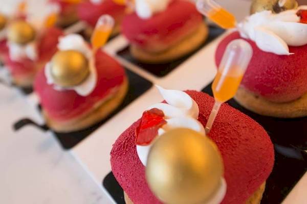 Mehrere rote Desserts mit goldenen Kugeln und orangefarbenen Stäbchen auf einem weißen Teller.