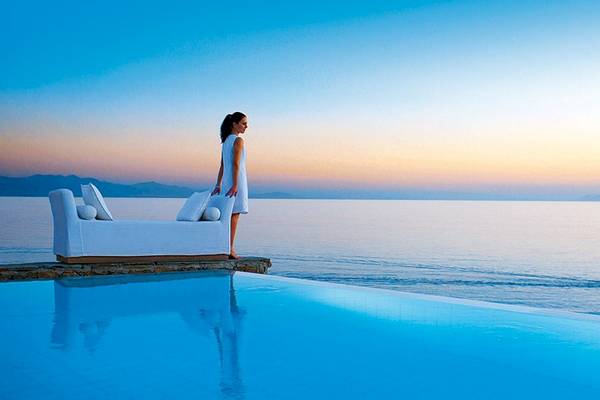 Eine Frau in einem weißen Kleid steht am Rand eines Swimmingpools und genießt den Blick auf den Sonnenuntergang.
