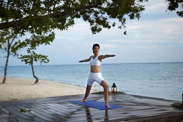 Frau in weißer Yoga-Kleidung praktiziert Yoga auf einer Holzplattform am Strand.