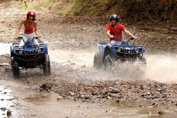 Zwei Leute auf Quads im Wasser, einer mit rotem Helm und roter Shirt, die andere mit rotem Helm und weißem Shirt.