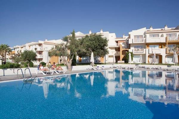 Zwei Personen entspannen am Pool des Blau Punta Reina Hotels, umgeben von Sonnenliegen und Bäumen.