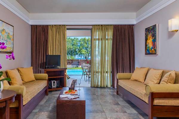 Das Wohnzimmer im Porto Platanias Beach Resort & Spa mit zwei Sofas, einem Tisch, einem Fernseher, einem Glastisch, einem Pool und einem Gemälde.