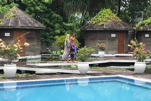Ein Paar steht neben einem Schwimmbad im Freien, umgeben von blumengeschmückten Hütten und Pflanzen.