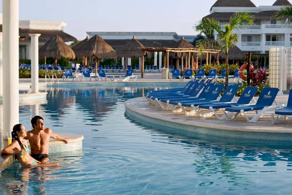 Paar im Pool des Grand Sunset Princess All Suites Resorts mit Liegestühlen und tropischer Umgebung
