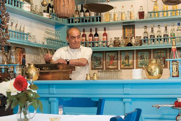 Ein Barkeeper des Grecotel Creta Palace steht hinter einer blauen Bar und bedient eine Kupfer-Spirituosenflasche.