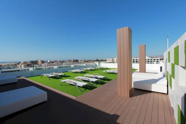 Dachterrasse des Caserio Hotels mit weißen Liegen auf grünem Kunstrasen, umgeben von Gebäuden.