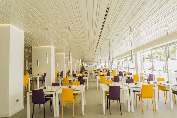 Innenansicht des Caserio Hotels Restaurant mit weißen Tischen, gelben und lila Stühlen, weißer Decke und großen Fenstern.