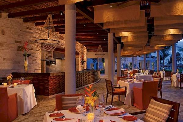 Innenbereich des Secrets Akumal Riviera Maya Hotels mit rustikalem Design, Säulen, Beleuchtung und gedeckten Tischen.