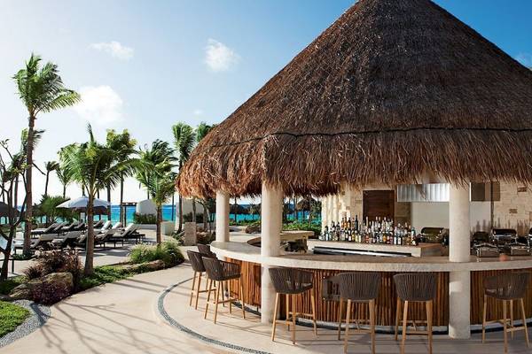 Eine Bar mit Strohdach im Secrets Akumal Riviera Maya Hotel, umgeben von Palmen und Sonnenliegen.