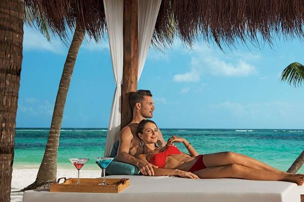 Paar im roten Bikini und Anzug entspannt auf einem Strandbett unter einem Sonnenschirm, mit Blick auf das Meer, Palmen und Cocktails.