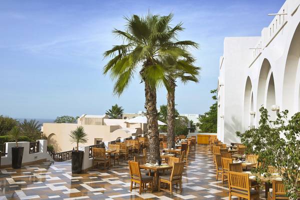 Terrasse des Cove Rotana Resorts mit braunen Holztischen und Stühlen unter zwei Palmen.