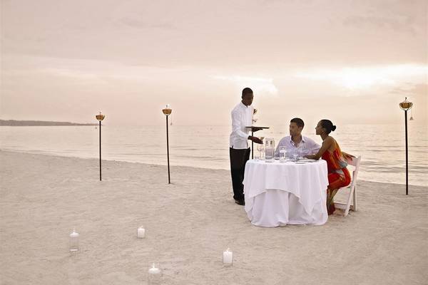 Eine Paar genießt ein Essen am Strand mit einem Kellner der eine Flasche Champagner serviert.