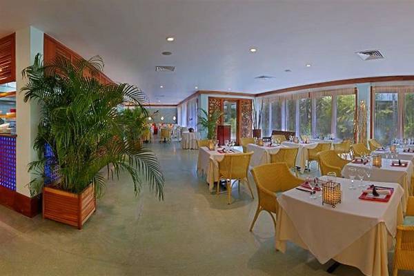 Innenansicht des Restaurants im Couples Negril Hotel mit gelben Stühlen und Tischen, einer großen Pflanze und Panoramafenstern.