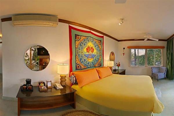 Schlafzimmer im Couples Negril Hotel mit gelbem Bett, orangenen Kissen und farbigem Wandteppich.