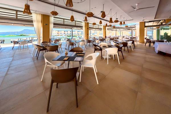 Innenansicht des Restaurants im Kiani Beach Resort mit vielen Tischen und Stühlen, großen Fenstern und Blick auf das Meer.