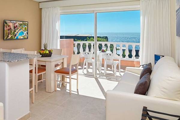 Balkon mit Meerblick im Aparthotel Allegro Isora mit Tisch, Stühlen, Fernseher und Obstkorb.