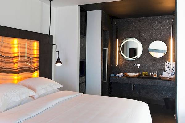 Schlafzimmer im Myconian Avaton Resort mit beleuchtetem Kopfteil und Badezimmer im Hintergrund.