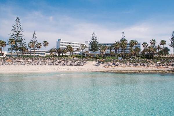 Außenansicht des Nissi Beach Resorts mit weißem Gebäude, Palmen und klarem Wasser.