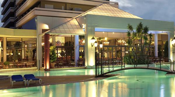 Der Außenpool des Dionysos Hotels bei Nacht mit einer Brücke und Lounge-Stuhl am Pool.
