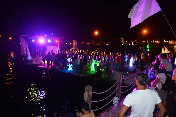Menschenmenge bei einer Nachtveranstaltung am Wasser, mit bunt beleuchteten Tischen und Flaggen