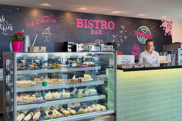 Innenansicht der Bistro Bar im H10 Rubicon Palace mit einer Wandtafel, die 'Bistro Bar' und 'ice cream' beschriftet ist, einer Verkäuferin und einer Vitrine voller Kuchen und Sandwiches.
