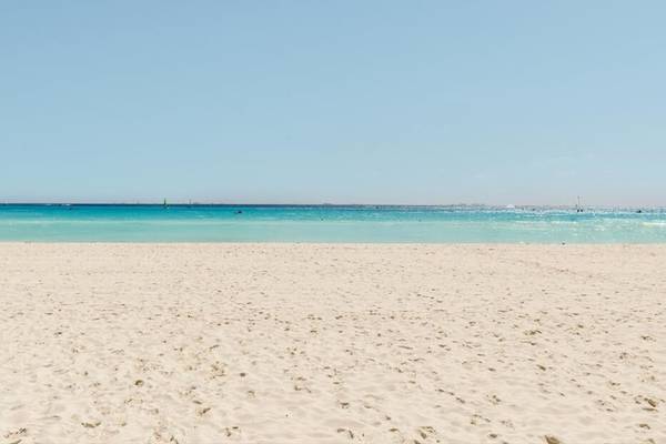 Foto von einem weißen Sandstrand und türkisfarbenem Wasser, mit einem blauen Himmel.