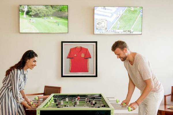 Gäste im Allegro Playacar Hotel spielen Tischfußball vor einem Fußballtrikot und Fernsehern an der Wand.